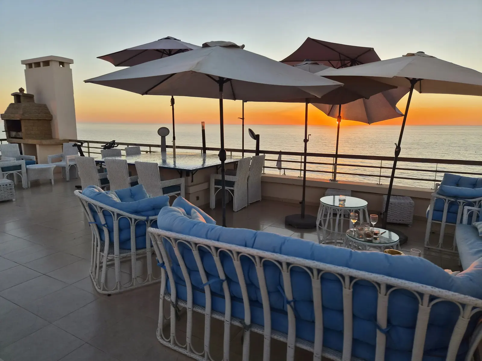 Grande terrasse du penthouse au coucher du soleil : salon d’extérieur aux coussins bleus, parasols, barbecue et océan Atlantique en fond